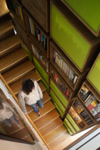 Photo from above a wood staircase looking down at a woman walking down the stairs. There is a mirror and handrail on the left of the image, with a green wood bookshelf on the right.