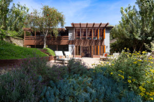 View of Back of House from Backyard. Includes flowers and trees surrounding a stone gathering circle with cushioned seats.