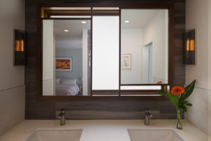 View of a modern bathroom counter. Includes a marble countertop. a black tile back, and two mirrors reflecting views of the bedroom and the hallway.