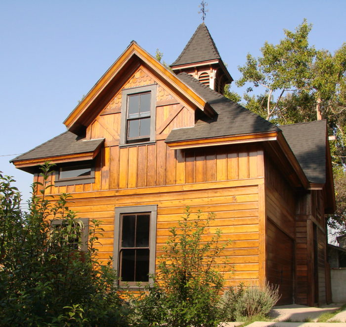Photo of the exterior of a modern house. Includes a wooden facade and black shingled roof, a stone slab front driveway, and surrounding foliage and trees.