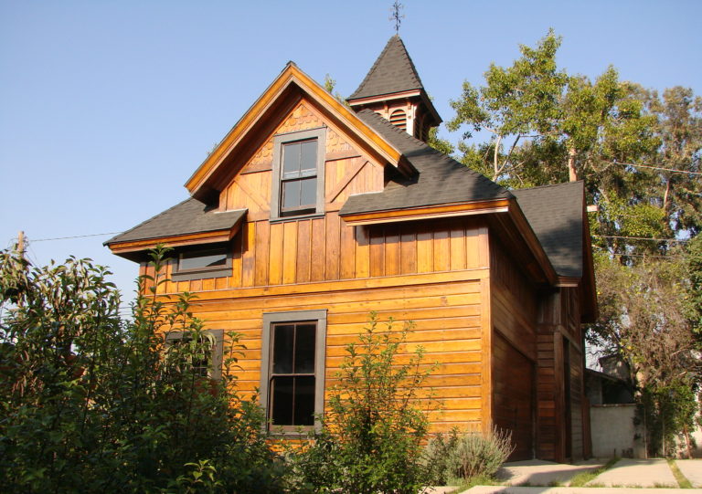 Photo of the exterior of a modern house. Includes a wooden facade and black shingled roof, a stone slab front driveway, and surrounding foliage and trees.