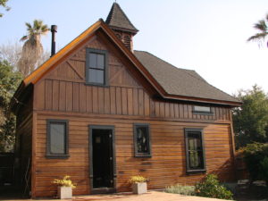 Photo of the exterior of a modern house. Includes a wooden facade and black shingled roof, a wood patio, and surrounding foliage and trees.