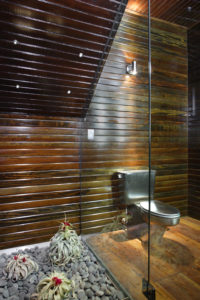 Photo of modern bathroom with wooden walls, floors, and ceiling. Includes a stainless steel toilet, a rock garden, and a glass partition separating the two.