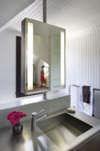 Photo of a modern bathroom with white wooden walls and ceiling. Includes a free-standing mirror and a stainless steel countertop against a pony tile wall.