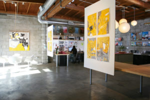 Interior photo of the Tracy A. Stone Architects office. Features a concrete floor and stone brick wall, hanging lights and posters, and a separate sitting area away from the work desks.