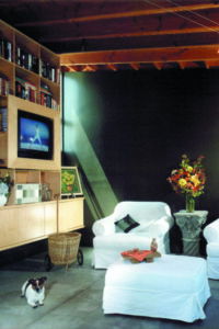 Photo of a living room for a modern house unit. Features a white chair and footrest, a wooden mantle with built-in television, and dark green walls with concrete floors.