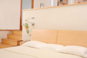 View of bed within modern bedroom. Includes a wooden staircase, white plaster walls, and an overhead bedside lamp.