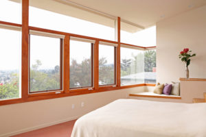 View of a bedroom in a modern house. Includes white plaster walls, multiple glass windows across from the bed, and a place to sit built into the wall in the corner.