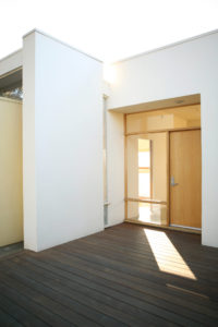 View of front entryway to a modern house. Shows a wooden floor, white stucco walls, and a wooden door surrounded by glass windows.