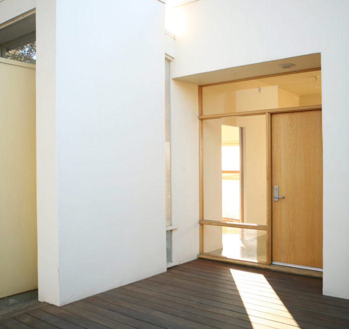 View of front entryway to a modern house. Shows a wooden floor, white stucco walls, and a wooden door surrounded by glass windows.