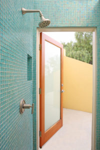 View of an outdoor shower for a modern home. Includes blue tile walls and an opaque glass wooden door.