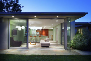 Photo of a modern house interior from outside. Includes a kitchen in the back with wooden cabinetry and a bar-height island, and a living room in the foreground with a sofa and mounted television.