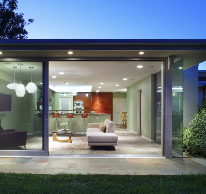 Photo of a modern house interior from outside. Includes a kitchen in the back with wooden cabinetry and a bar-height island, and a living room in the foreground with a sofa and mounted television.