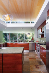 View of the kitchen and backyard of a modern apartment. Shows the wooden cabinets and drawers, marble countertops, stainless steel appliances, and large floor to ceiling windows.