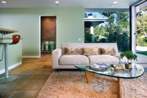 Photo of living room of a modern house. Features tile floors, green painted walls, floor-to-ceiling windows on the right side, and a kitchen island on the left side.