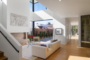 Photo of the living room of the Keniston Residence. Features white walls, a wine cellar to the right, a living room with an open air second floor above, and a open sliding glass door in the background.