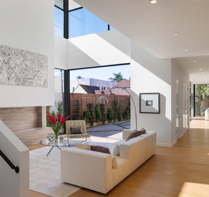 Photo of the living room of the Keniston Residence. Features white walls, a wine cellar to the right, a living room with an open air second floor above, and a open sliding glass door in the background.