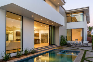 Photo of backyard and pool of a modern house. Includes white walls, turfstone pavers. and a look into the kitchen and living room through an open sliding glass door.