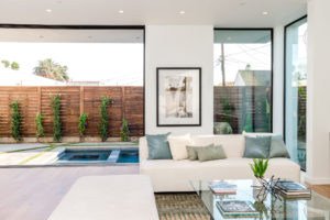 Photo of living room and backyard within a modern house. Includes wood floors, white stucco walls and ceiling, floor to ceiling windows and sliding doors, and a view at the backyard pool.