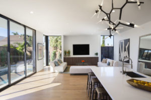 Photo of living room and kitchen within a modern house. Includes wood floors, white stucco walls and ceiling, a kitchen island, and floor to ceiling windows and sliding doors.