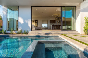 Photo of backyard and pool within a modern house. The pool rests in the foreground of the photo, while the background includes wood floors, white stucco walls and ceiling, floor to ceiling windows and sliding doors.