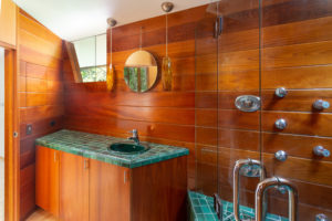 Photo of a bathroom within a modern house. Features a wooden wall with a circular mirror, blue tiles covering the countertop and shower, and a window to the outside in the top left.