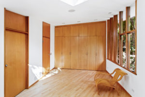 Photo of bedroom within modern residence. Features wooden floors, dressers, and doors, white stucco walls, and a single wooden chair beneath multiple windows.