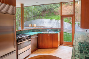 Photo of kitchen within a modern house. Features wooden floors and cabinets, blue tile countertops, a fridge and oven, and floor-to-ceiling windows.