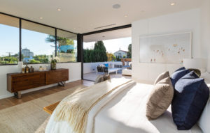 View of modern bedroom with an open sliding glass door that leads out onto an outdoor sitting area. Includes white plaster walls, a wooden dresser across from the bed, and large windows behind the dresser.