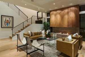 View of a living room within a modern house. Includes wooden floors, white stucco walls and ceiling, a fireplace and hearth made of wood and stone, and a staircase in the background with black railings.