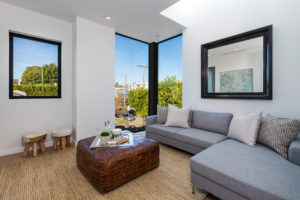 View of modern Living Room with Corner Windows. Includes white plaster walls, a mirror hanging above the "L" shaped couch, and a large footrest cushion that is holding various items.