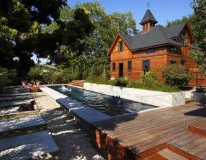 A vintage Los Angeles carriage house with a swimming pool addition. Includes a wooden facade and black shingled roof, a swimming pool, and a backyard that has both wooden patio space and stone slabs embedded between grass.