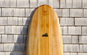 Close up picture of a wooden surfboard leaning against a wooden shingle wall.