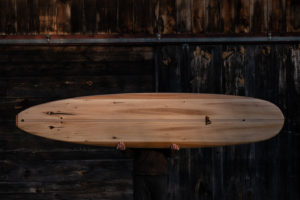 Photo of a person holding a wooden surfboard in front of their face. They are holding the board horizontally while standing against a dark wooden background.