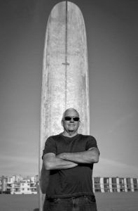 Black-and-white photo of a man standing in front of a surfboard with his arms crossed. He is wearing a dark colored shirt and slightly smiling.