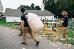 Photo of a man carrying a bag over his shoulder as he walks up a path. There are other people in the background working on crafting wooden surfboards.