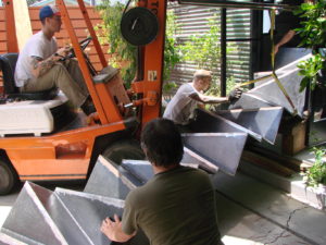 Photos showing multiple men and a forklift being used to move a modern staircase indoors. The stair is tilted on its side.