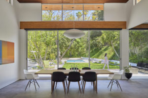 Photo of a dining room and backyard of a modern house. The kitchen features a wooden table with eight chairs on dark tiles and white stucco walls, with floor-to-ceiling windows revealing a backyard featuring a pool, trees, and grass.