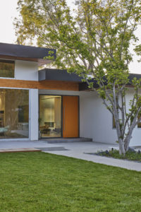 Rendering of the backyard and back facade of a modern house. Includes a concrete patio and walkway, a floor to ceiling glass sliding door going into the living room, and a grass yard.