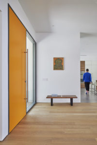 View of the interior entryway of a modern house. Shows the wood floors and white stucco walls, and a large orange door to the left of the image with a vertical metal handlebar on the right.