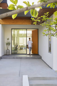 View of an exterior entryway of a modern house. Features a concrete staircase and patio leading to a large orange door and large glass window, all sitting under a wooden roof overhang.