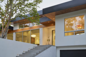 Photo of the entryway of a modern house. Concrete stairs lead up to an exterior facade that includes white and wooded walls, multiple large windows, and an overhanging black roof.