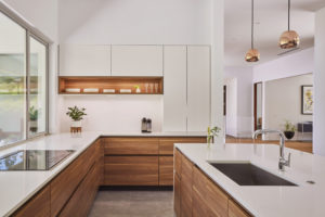 Photo of a kitchen within a modern house. Features white marble countertops. natural and white painted wood cabinetry, large windows to the left, and an island to the right with two hanging lights.