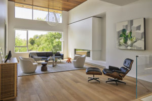 Photo of a modern living room for a modern house. Includes a wooden floor and white stucco walls, a built-in fireplace on the right wall, and multiple windows on the back wall.