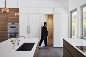 Kitchen of a modern unit that features wooden cabinets, stainless steel appliances, gray marble floor, and windows on the right. A man open a hidden door nestled between white cabinets to reveal a washer and dryer room,