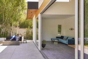 Photo of a patio and living room for a modern house. The right side shows the living room through an open floor-to-ceiling sliding glass door, while the left side shows a couple sitting in front of an outdoor fireplace on a sofa.