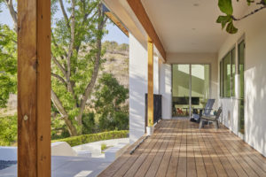 Photo of a porch for a modern house. Features a wooden floor, white stucco walls and ceiling, wooden beams and columns, and a black railing.