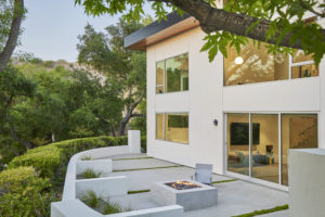 Photo of the backyard of an modern house. Shows large concrete pavers, a concrete firepit, and the white exterior walls of the house with multiple large glass windows.