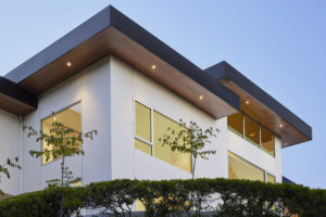 Photo of the roof of a modern house. Includes white and wooded walls, multiple large windows, an overhanging black roof, and a line of bushes in the foreground.