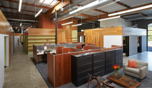 Rendered view of primary work space within a modern office. Includes wood wall panels against a green wall, multiple wooden cubicles, and a small lounge seating area.